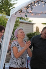 A group of people are standing together under a white canopy adorned with string lights. One person is positioned in the center with closed eyes and clasped hands, possibly in a moment of reflection or prayer. A lush garden and a house are visible in the background.