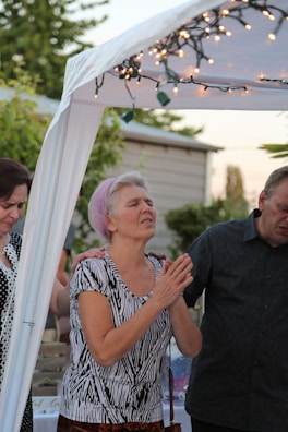 A group of people are standing together under a white canopy adorned with string lights. One person is positioned in the center with closed eyes and clasped hands, possibly in a moment of reflection or prayer. A lush garden and a house are visible in the background.