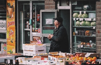 A street vendor stands near the entrance of a small store, surrounded by an array of fresh produce displayed in boxes. Cabbages, oranges, and various fruits are prominently placed in front of the shop. The exterior of the store has a combination of brick and glass, with posters advertising different products. The man is wearing a dark jacket and a cap, seemingly waiting for customers.