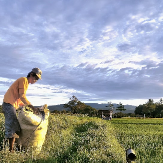 A smiling farmer holding a sack of fertiliser in a lush green field under a bright sky.