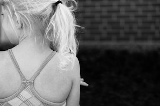 Young girl with light blonde hair tied in a ponytail, wearing a plaid-patterned strappy dress or swimsuit. Her back is turned to the camera, with a brick wall in the background.