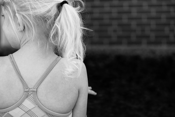 Young girl with light blonde hair tied in a ponytail, wearing a plaid-patterned strappy dress or swimsuit. Her back is turned to the camera, with a brick wall in the background.