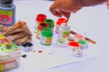 A hand holding a paintbrush is seen dipping into small containers of paint. The scene includes several jars of vibrant acrylic colors such as red, green, and orange, placed on a white surface. There are paint stains on the paper beneath them, and a cloth is positioned nearby, likely used for cleaning. The overall setup suggests an art project or painting in progress.