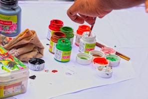 A hand holding a paintbrush is seen dipping into small containers of paint. The scene includes several jars of vibrant acrylic colors such as red, green, and orange, placed on a white surface. There are paint stains on the paper beneath them, and a cloth is positioned nearby, likely used for cleaning. The overall setup suggests an art project or painting in progress.
