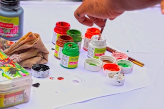 A hand holding a paintbrush is seen dipping into small containers of paint. The scene includes several jars of vibrant acrylic colors such as red, green, and orange, placed on a white surface. There are paint stains on the paper beneath them, and a cloth is positioned nearby, likely used for cleaning. The overall setup suggests an art project or painting in progress.
