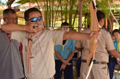A man in a beige shirt is aiming a wooden recurve bow with an arrow drawn back, surrounded by other people, including a group in colorful uniforms. There is a lush green background with bamboo plants.