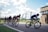 Cyclists are racing along a track on a sunny day. A group of riders, wearing cycling gear and helmets, are focused and concentrated as they navigate a curve. The setting appears to be a cycling event with flags lining the road and a wooden building in the background. A person is standing nearby, possibly taking photos or observing the race.