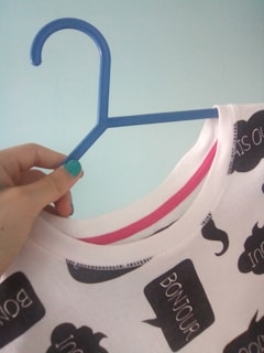 A hand with painted nails is holding a white shirt with speech bubble patterns, some of which contain the word 'BONJOUR'. The shirt is on a blue plastic hanger against a light blue background.