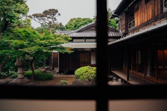 A traditional wooden Japanese house with sliding doors and a tiled roof is surrounded by a lush garden featuring manicured plants and trees. The foreground reveals a serene courtyard with greenery. The image is viewed through a window, framed by wooden beams.