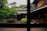 A traditional wooden Japanese house with sliding doors and a tiled roof is surrounded by a lush garden featuring manicured plants and trees. The foreground reveals a serene courtyard with greenery. The image is viewed through a window, framed by wooden beams.