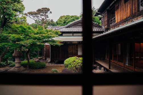 A traditional wooden Japanese house with sliding doors and a tiled roof is surrounded by a lush garden featuring manicured plants and trees. The foreground reveals a serene courtyard with greenery. The image is viewed through a window, framed by wooden beams.