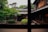 A traditional wooden Japanese house with sliding doors and a tiled roof is surrounded by a lush garden featuring manicured plants and trees. The foreground reveals a serene courtyard with greenery. The image is viewed through a window, framed by wooden beams.