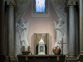 A chapel interior with a central altar flanked by two painted angels on the walls. The altar features an ornate tabernacle, a crucifix, and a red sanctuary lamp. The setting includes classical architectural elements like columns and drapery.