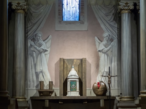 A chapel interior with a central altar flanked by two painted angels on the walls. The altar features an ornate tabernacle, a crucifix, and a red sanctuary lamp. The setting includes classical architectural elements like columns and drapery.