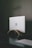 A silver laptop with an Apple logo partially closed, resting on a curved wooden stand against a dark textured wall. A power cable is plugged into the left side of the laptop. The surface it is placed on is black, creating a minimalist and modern aesthetic.