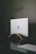 A silver laptop with an Apple logo partially closed, resting on a curved wooden stand against a dark textured wall. A power cable is plugged into the left side of the laptop. The surface it is placed on is black, creating a minimalist and modern aesthetic.