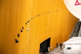 A tall, curved yellow wall is adorned with numerous security cameras arranged in a precise diagonal line. Below, several people are walking along a pathway beside a series of white tents or structures labeled as 'Press Desk'. A staircase on the right side leads away from the scene.