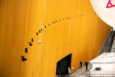 A tall, curved yellow wall is adorned with numerous security cameras arranged in a precise diagonal line. Below, several people are walking along a pathway beside a series of white tents or structures labeled as 'Press Desk'. A staircase on the right side leads away from the scene.
