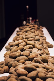 A large pile of potatoes is spread across a long, narrow table with a dark background. The tips of various spice jars can be seen blurred in the distance, suggesting a culinary or kitchen setting.