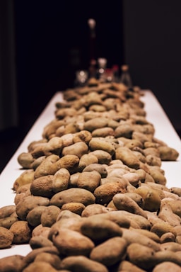 A large pile of potatoes is spread across a long, narrow table with a dark background. The tips of various spice jars can be seen blurred in the distance, suggesting a culinary or kitchen setting.