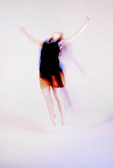 A person appears to be in a pose of mid-air jump or dance, with arms extended upward. The image conveys a sense of motion and movement through the slightly blurred outline of the figure.