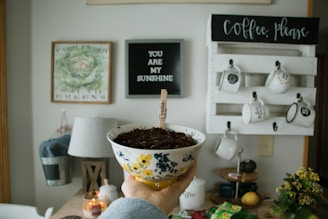 A hand holds a decorative bowl filled with soil, with a popsicle stick sign labeled 'lemon balm'. The background features a cozy kitchen setting with a lamp, plants, a candle, and a wooden wall rack displaying mugs. Two framed wall hangings are visible, one with a botanical illustration and another with the text 'YOU ARE MY SUNSHINE'.