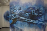 A large mural painted on a wall depicting an abstract aerial view of a cityscape. The artwork features a variety of buildings and streets, rendered in a blend of blues, blacks, and whites. There is a noticeable drip effect, creating a sense of movement or rain. A bridge with towers can be seen, adding to the urban landscape.