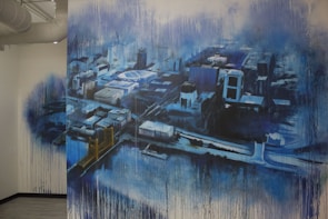 A large mural painted on a wall depicting an abstract aerial view of a cityscape. The artwork features a variety of buildings and streets, rendered in a blend of blues, blacks, and whites. There is a noticeable drip effect, creating a sense of movement or rain. A bridge with towers can be seen, adding to the urban landscape.