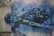 A large mural painted on a wall depicting an abstract aerial view of a cityscape. The artwork features a variety of buildings and streets, rendered in a blend of blues, blacks, and whites. There is a noticeable drip effect, creating a sense of movement or rain. A bridge with towers can be seen, adding to the urban landscape.