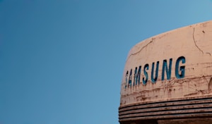 A weathered, vintage sign with the word 'SAMSUNG' in faded blue letters against a heavily worn, off-white background, set against a clear blue sky.