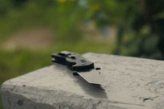 A folding knife is placed on an outdoor concrete surface with a blurred green background.