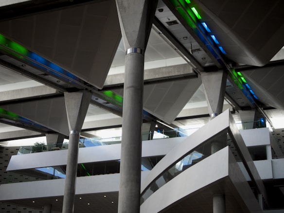 A modern architectural interior with large, exposed concrete support columns and angular white walls. The ceiling features colorful lighting elements with strips of green and blue lights. Glass railings are present, adding to the open and airy feel of the space.