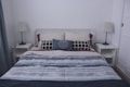 A neatly made bed with a patterned comforter featuring various shades of gray and blue, accompanied by two white pillows and a central decorative pillow with a geometric design. Each side of the bed has a small white nightstand, each topped with a lamp that has a pleated white shade. The walls are plain, enhancing the minimalist aesthetic.