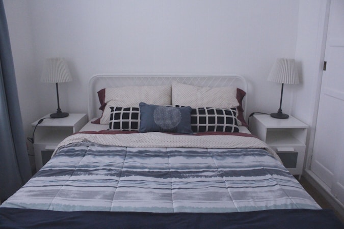 A neatly made bed with a patterned comforter featuring various shades of gray and blue, accompanied by two white pillows and a central decorative pillow with a geometric design. Each side of the bed has a small white nightstand, each topped with a lamp that has a pleated white shade. The walls are plain, enhancing the minimalist aesthetic.