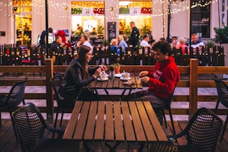 A cozy outdoor café setting at night with string lights adding a warm ambiance. Two people are sitting at a wooden table engaged in conversation, each with a drink. The background is bustling with other patrons seated at tables, contributing to a lively atmosphere.
