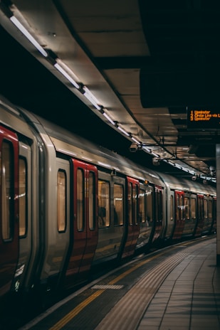 A subway train is stationed at a platform in an underground transit station. The train features red and white colors, with sliding doors visible. Overhead lights illuminate the area, while a digital sign displays upcoming train destinations in bright yellow text.