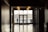 A silhouette of a person leaning on a railing outside near water is visible through large glass doors at the end of a dimly lit hallway. The floor is shiny, reflecting overhead lights. A sign for restrooms is mounted on the wall to the right.