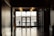 A silhouette of a person leaning on a railing outside near water is visible through large glass doors at the end of a dimly lit hallway. The floor is shiny, reflecting overhead lights. A sign for restrooms is mounted on the wall to the right.