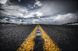 An asphalt road stretches into the distance, with a vibrant yellow line running down the center. The sky above is filled with dramatic clouds, casting varied lighting on the scene. The road appears wet, with reflective patches of water. Sparse vegetation is visible on the horizon.