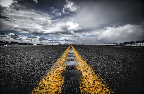 An asphalt road stretches into the distance, with a vibrant yellow line running down the center. The sky above is filled with dramatic clouds, casting varied lighting on the scene. The road appears wet, with reflective patches of water. Sparse vegetation is visible on the horizon.