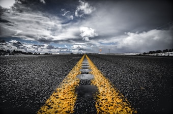 An asphalt road stretches into the distance, with a vibrant yellow line running down the center. The sky above is filled with dramatic clouds, casting varied lighting on the scene. The road appears wet, with reflective patches of water. Sparse vegetation is visible on the horizon.