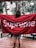 A person stands on a wooden deck facing away, with a large red towel draped over their shoulders. The towel prominently features the word 'Supreme' in white letters. In the background, there are tall pine trees, creating a natural, forest-like setting.