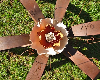 A metal sculpture resembling a large flower with rusted metal petals and intricate wire detailing is set against a backdrop of green grass. The inner part of the sculpture has layered textures and varying shades, creating a dynamic visual contrast.