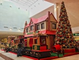 A large, decorated Christmas tree stands next to a festive train ride inside a shopping mall. The train and nearby house-like structure have a classic red and green holiday theme, adorned with garlands and wreaths. Shoppers, including adults and children, are interacting with the display.