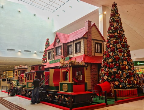 A large, decorated Christmas tree stands next to a festive train ride inside a shopping mall. The train and nearby house-like structure have a classic red and green holiday theme, adorned with garlands and wreaths. Shoppers, including adults and children, are interacting with the display.