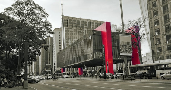 A modern urban street scene features a prominent building with distinct red columns. The architecture has a sleek and contemporary design with glass and concrete elements. Vehicles, including cars and a bus, are visible on the busy road. Trees and various billboards add to the cityscape ambiance.