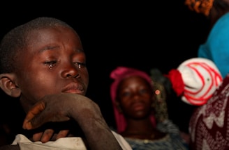 A close-up of a young boy with a tear on his cheek, eyes closed, displaying a contemplative or sad expression. In the background, partially blurred figures include another child wearing a pink headscarf and an adult dressed in vibrant clothing. The scene takes place against a dark backdrop, suggesting nighttime or a dimly lit environment.
