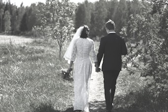 A couple is walking hand in hand along a grassy path, surrounded by lush greenery and trees. The woman is wearing a white gown with a veil, holding a bouquet, and the man is in a dark suit. The setting appears serene and natural, with a soft, muted color palette.