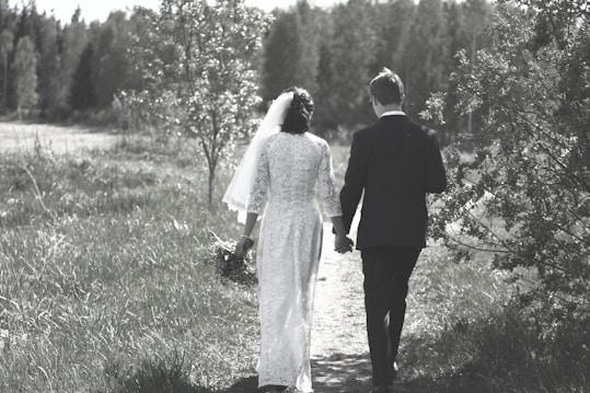 A couple is walking hand in hand along a grassy path, surrounded by lush greenery and trees. The woman is wearing a white gown with a veil, holding a bouquet, and the man is in a dark suit. The setting appears serene and natural, with a soft, muted color palette.