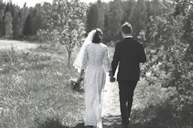 A couple is walking hand in hand along a grassy path, surrounded by lush greenery and trees. The woman is wearing a white gown with a veil, holding a bouquet, and the man is in a dark suit. The setting appears serene and natural, with a soft, muted color palette.